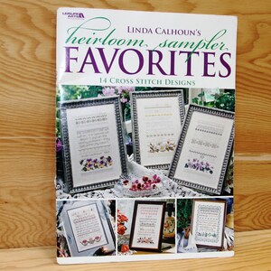 May include: Book cover for "Heirloom Sampler Favorites" by Linda Calhoun. The cover features 14 cross stitch designs, with a floral theme and a quote about the beauty of nature. The book is published by Leisure Arts.