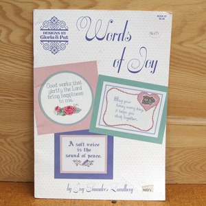 May include: A vintage craft book titled "Words of Joy" by Gloria & Pat. The cover features cross-stitch designs with inspirational quotes and floral motifs in pink, blue, and green. The book is displayed on a wooden surface.