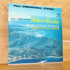 May include: A vintage vinyl record album cover featuring a scenic aerial view of a Hawaiian island with the text "Island Paradise" and "WEBLEY EDWARDS PRESENTS" in the upper left corner. The album cover also includes the text "EXCITING, ON-THE-SPOT RECORDINGS: THE AUTHENTIC SOUNDS AND MUSIC OF HAWAII, THE 50TH STATE."