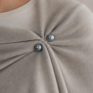 Könnte beinhalten: Ein hellgraues Stricktuch mit einer glatten, feinmaschigen Textur. Das Kleidungsstück ist an der Schulter mit zwei runden, dunkelgrauen perlenartigen Knöpfen befestigt, die weiche, drapierte Falten über der Brust und Schulter erzeugen.