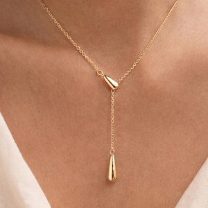 Könnte beinhalten: Eine zarte, goldfarbene Halskette mit einem Tropfenanhänger. Die Halskette hat eine feine Kette mit einem kleinen tropfenförmigen Akzent in der Nähe des Verschlusses. Der Hauptanhänger ist ein größerer, polierter Tropfen, der an einer längeren Kette hängt.