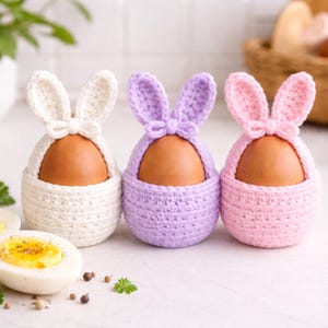 May include: Three crocheted egg holders in white, lavender, and pink, each shaped like a bunny with upright ears and a bow. Brown eggs sit inside the holders. Sliced hard-boiled eggs and parsley are in the foreground, suggesting an Easter theme.