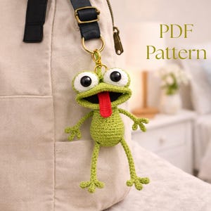 May include: A green crocheted frog keychain with large white-rimmed eyes and a red tongue. The frog is attached to a gold-colored keyring and hangs from a beige bag. The text "PDF Pattern" is in the upper right corner.