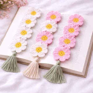 Puede incluir: Marcadores de ganchillo con forma de margarita en blanco y rosa con centros amarillos y borlas verdes o crema. Los marcadores están colocados en las páginas de un libro abierto, con un fondo suave y borroso.