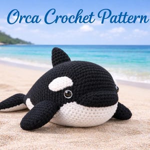 May include: A crocheted orca whale toy in black and white, lying on a sandy beach. The toy has a dorsal fin, flippers, and white patches. The text "Orca Crochet Pattern" is written in blue cursive above the whale.