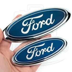 Pode incluir: Dois emblemas ovais da Ford. Apresentam a palavra "Ford" em prata sobre um fundo azul escuro, emoldurados por uma borda cromada. Os emblemas são segurados em uma mão.