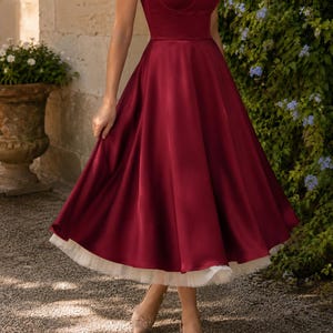 May include: A deep burgundy satin midi dress featuring a sweetheart neckline, thin spaghetti straps, and a fitted bodice. The skirt is full and flared, showcasing a layered white tulle petticoat underneath that extends slightly below the hemline.