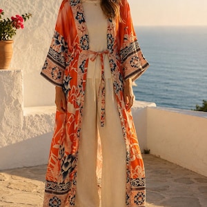 Può includere: Un lungo kimono aperto sul davanti in una tonalità arancione vivace, caratterizzato da un dettagliato motivo floreale e geometrico nei toni del blu, bianco e nero. Il capo presenta maniche ampie, una cintura da annodare e un orlo lungo.