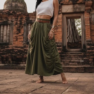 May include: An olive green skirt with gathered detailing and a brown belt is paired with a white crop top. The skirt has a flowing, bohemian style. The outfit is worn in front of a weathered brick building.