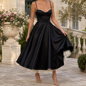 May include: A black midi dress featuring a structured bodice with thin spaghetti straps and a sweetheart neckline trimmed with small white beads. The full, flared skirt has a visible white tulle petticoat layer underneath the hemline.