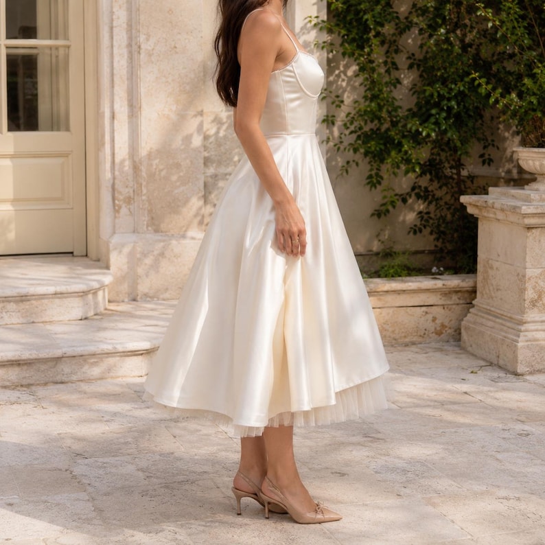 May include: A cream-colored midi dress featuring a structured bodice with thin shoulder straps and a flared skirt. The skirt has a smooth satin-like outer layer that falls to mid-calf, revealing a delicate tulle hemline underneath the dress.