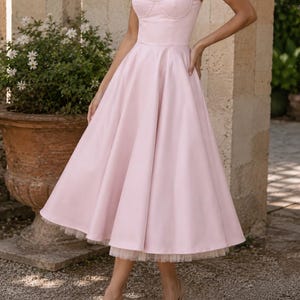 May include: A light pink midi dress featuring a fitted bodice with a sweetheart neckline, thin spaghetti straps, and a full, flared skirt. The hemline reveals a layer of delicate white tulle underneath, creating a structured and voluminous silhouette.