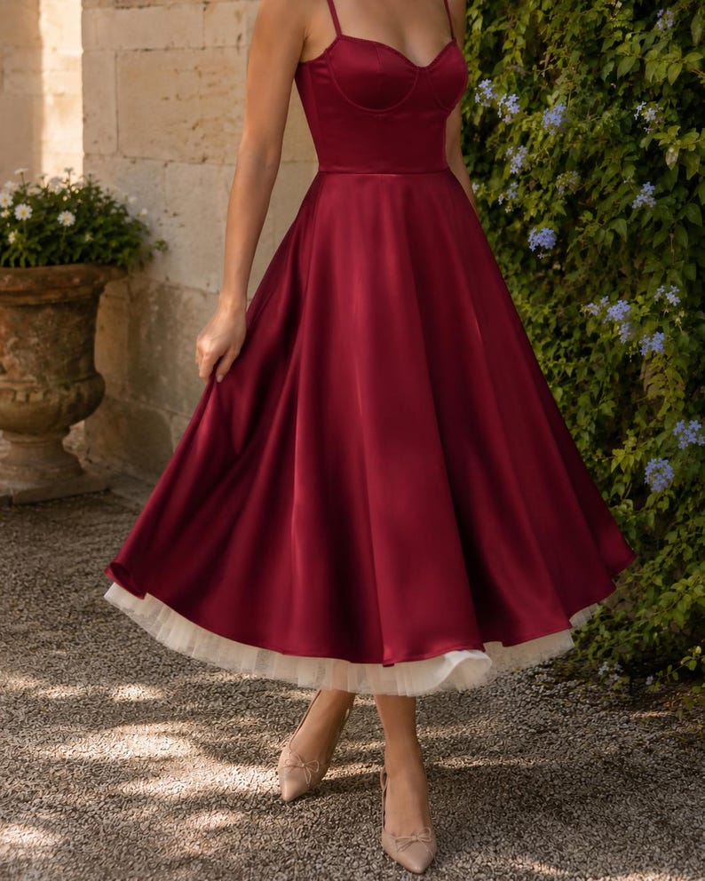 May include: A deep burgundy satin midi dress featuring a sweetheart neckline, thin spaghetti straps, and a fitted bodice. The skirt is full and flared, showcasing a layered white tulle petticoat underneath that extends slightly below the hemline.