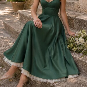 May include: A dark green midi dress featuring a structured sweetheart neckline, thin spaghetti straps, and a full skirt with a visible white tulle petticoat hem. The dress is paired with neutral-toned pointed-toe heels.