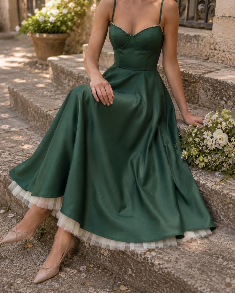 May include: A dark green midi dress featuring a structured sweetheart neckline, thin spaghetti straps, and a full skirt with a visible white tulle petticoat hem. The dress is paired with neutral-toned pointed-toe heels.