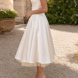 May include: A tea-length ivory satin dress featuring a fitted bodice with thin spaghetti straps and a full, flared skirt. The hemline reveals a layered tulle petticoat underneath, adding volume to the silhouette of this sleeveless garment.