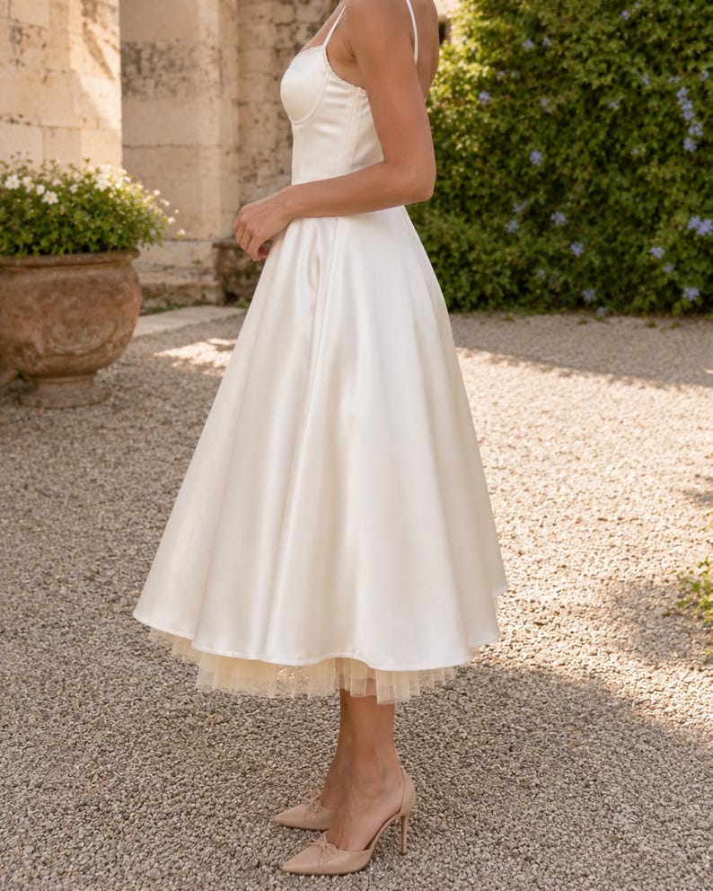 May include: A tea-length ivory satin dress featuring a fitted bodice with thin spaghetti straps and a full, flared skirt. The hemline reveals a layered tulle petticoat underneath, adding volume to the silhouette of this sleeveless garment.