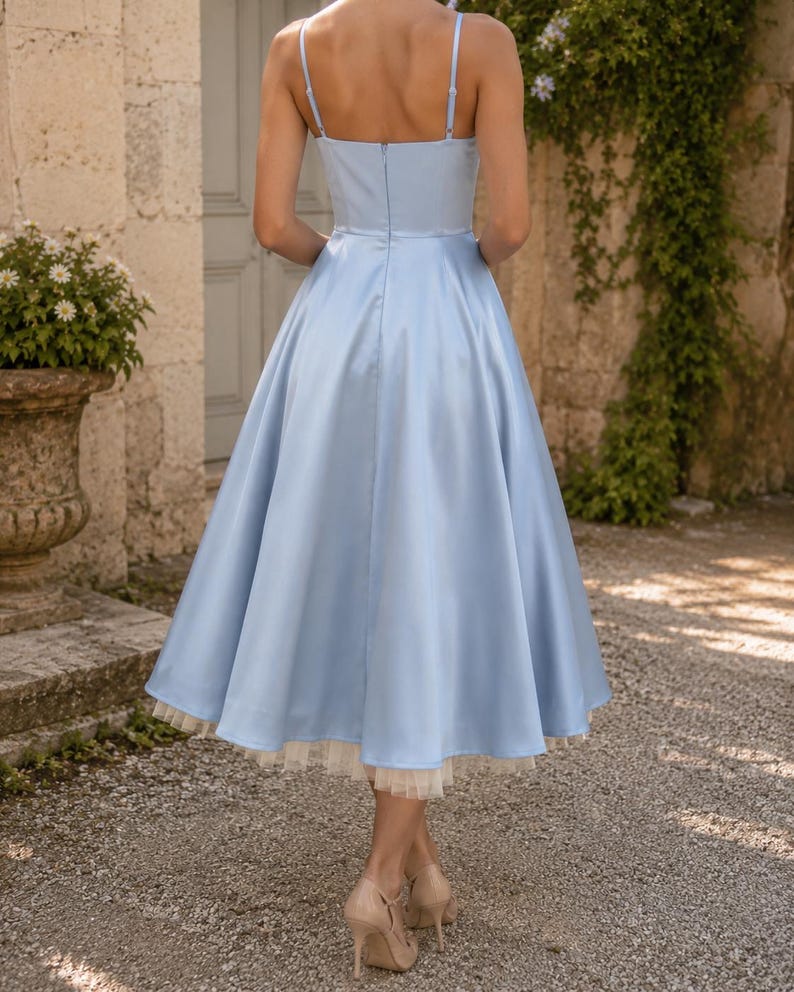 May include: A light blue midi dress featuring a fitted bodice with thin spaghetti straps, a flared skirt, and a visible cream-colored tulle petticoat hemline. The dress has a center back zipper closure and a smooth, satin-like fabric finish.
