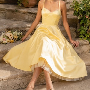 May include: A pale yellow satin midi dress featuring a sweetheart neckline, thin spaghetti straps, and a full, flared skirt. The dress includes a visible white tulle petticoat layer underneath the hemline, creating a structured and voluminous silhouette.