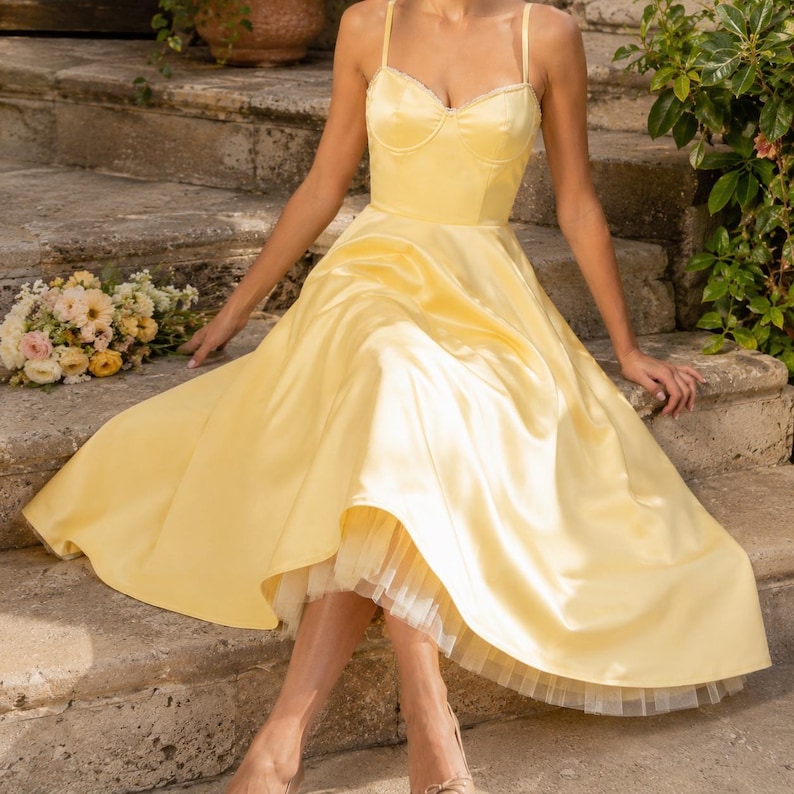 May include: A pale yellow satin midi dress featuring a sweetheart neckline, thin spaghetti straps, and a full, flared skirt. The dress includes a visible white tulle petticoat layer underneath the hemline, creating a structured and voluminous silhouette.