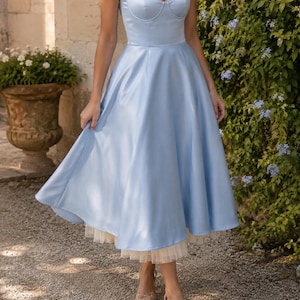 May include: A light blue midi dress featuring a structured sweetheart neckline, thin spaghetti straps, and a full, flared skirt. The garment includes a visible cream-colored tulle petticoat layer underneath the hemline, creating a layered effect.