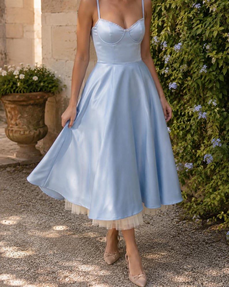 May include: A light blue midi dress featuring a structured sweetheart neckline, thin spaghetti straps, and a full, flared skirt. The garment includes a visible cream-colored tulle petticoat layer underneath the hemline, creating a layered effect.
