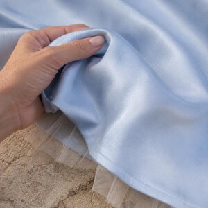 May include: A light blue satin fabric with a smooth, reflective surface, layered over a delicate white tulle underskirt. The fabric is gathered in soft folds, showcasing the sheen of the material and the contrast between the satin and tulle.