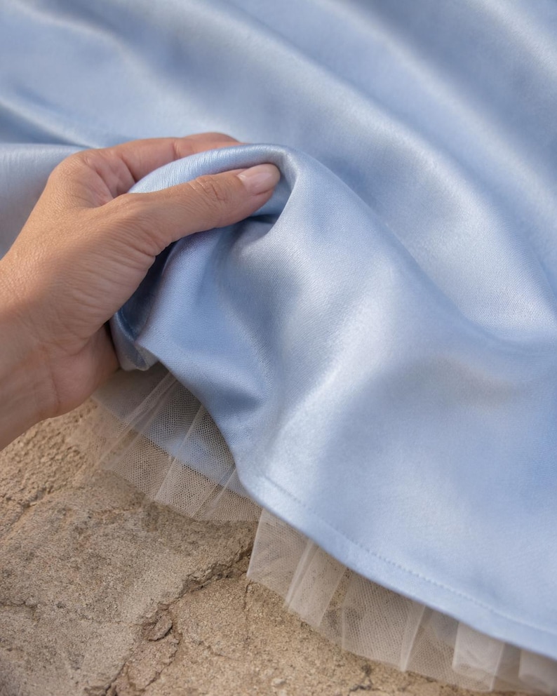 May include: A light blue satin fabric with a smooth, reflective surface, layered over a delicate white tulle underskirt. The fabric is gathered in soft folds, showcasing the sheen of the material and the contrast between the satin and tulle.