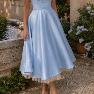 May include: A light blue midi dress featuring a sweetheart neckline, thin spaghetti straps, and a fitted bodice. The skirt is full and flared, with a visible cream-colored tulle petticoat layer extending slightly below the hemline of the dress.