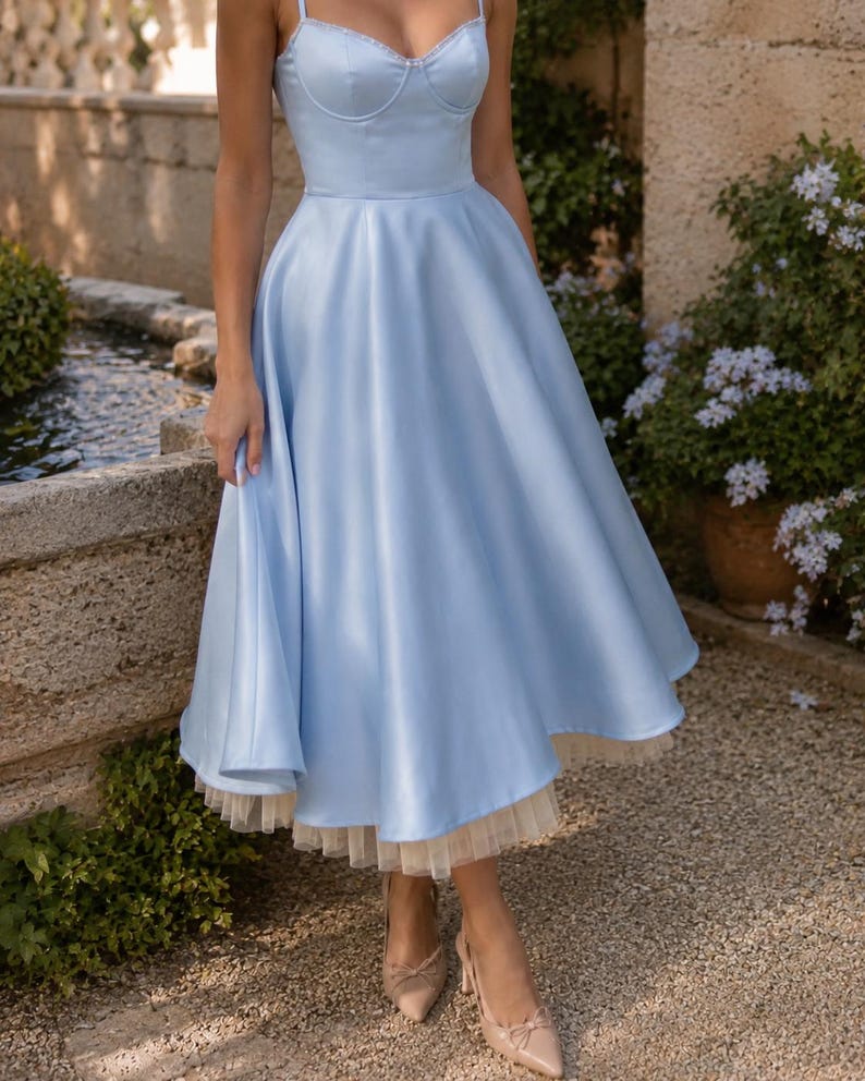 May include: A light blue midi dress featuring a sweetheart neckline, thin spaghetti straps, and a fitted bodice. The skirt is full and flared, with a visible cream-colored tulle petticoat layer extending slightly below the hemline of the dress.