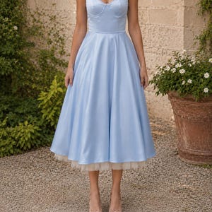 May include: A light blue midi dress with a structured sweetheart neckline, thin spaghetti straps, and a full, flared skirt. The hemline features a visible layer of cream-colored tulle petticoat underneath the satin-finish fabric of the dress.