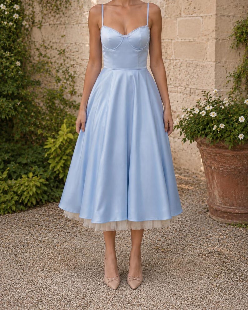 May include: A light blue midi dress with a structured sweetheart neckline, thin spaghetti straps, and a full, flared skirt. The hemline features a visible layer of cream-colored tulle petticoat underneath the satin-finish fabric of the dress.
