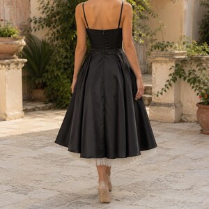 May include: A black midi-length dress with thin spaghetti straps and a fitted bodice. The skirt features a full, pleated silhouette that flares outward, revealing a white tulle petticoat layer underneath the hemline of the black fabric.