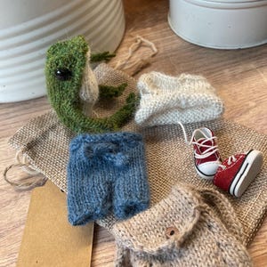 Può includere: Una collezione di articoli in miniatura lavorati a maglia, tra cui una rana verde, un maglione bianco, pantaloncini blu, uno zaino e scarpe da ginnastica rosse. Gli articoli sono disposti su un sacco di iuta, suggerendo un tema di avventura all'aperto o gioco.