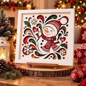May include: A square white-framed layered paper art piece featuring a central snowman wearing a red patterned hat and scarf with mittens. The design includes white snowflakes, green holly leaves, and red berries set against swirling red, green, and gold layers.