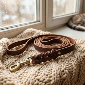 Könnte beinhalten: Eine aufgerollte braune Leder-Hundeleine mit einem messingfarbenen Karabinerhaken und einem geflochtenen Detail in der Nähe des Verschlusses. Die Leine liegt auf einer cremefarbenen Strickdecke in der Nähe eines Fensters.