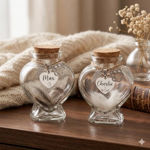 May include: Two heart-shaped glass keepsake jars with cork stoppers. Each jar has a silver charm with a paw print and wings, and a name tag that reads "Max" and "Charlie". The jars contain pet fur.