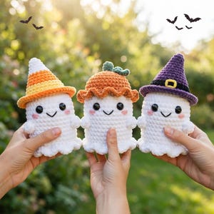 Può includere: Tre fantasmi bianchi all'uncinetto con occhi neri e guance rosa. Il fantasma a sinistra indossa un cappello a forma di caramella di mais gialla e arancione, quello centrale un cappello a zucca arancione e quello a destra un cappello da strega.