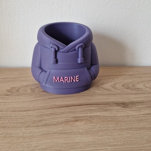 Könnte beinhalten: Ein violetter Blumentopf in Form eines Hoodies mit Kapuze, Kordelzug und Taschen. Das Wort "MARINE" ist in Rosa auf der Vorderseite aufgedruckt. Der Topf besteht aus einem festen Material und steht auf einer Holzoberfläche.