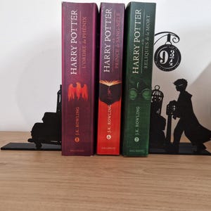 Peut inclure: Trois éditions françaises des livres "Harry Potter" sont placées entre des serre-livres en métal noir. Les dos des livres sont bordeaux, rouges et verts. Les serre-livres présentent une silhouette de la voie 9 3/4 et d'un sorcier.