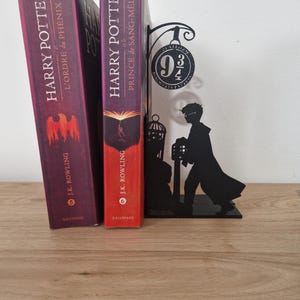 Op de afbeelding: Twee Franse edities van Harry Potter-boeken, "L'Ordre du Phénix" en "Prince de Sang-Mêlé", staan naast een zwarte metalen boekensteun van Perron 9 3/4. De boeken hebben bordeauxrode covers met gouden letters.