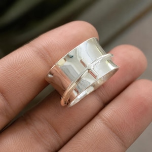 Peut inclure: Une large bague rotative en argent avec une bande polie à la surface lisse. Un fil fin de couleur or rose est enroulé autour du centre de la bande, formant une petite croix qui repose contre la surface métallique argentée réfléchissante.