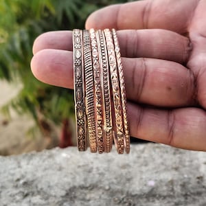 May include: A set of six copper-colored bangles with intricate embossed designs. Each bangle features a unique pattern, including floral, geometric, and textured elements. The bangles are displayed against a blurred background.