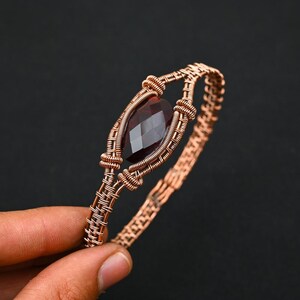 May include: Handmade copper wire bracelet with a faceted, dark red gemstone. The gemstone is set within a woven wire frame, and the bracelet band is also made from intricately woven copper wire. The design highlights a combination of natural materials and skilled craftsmanship.