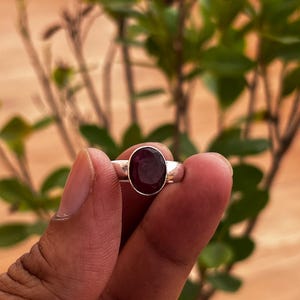 Puede incluir: Un anillo de plata con una piedra preciosa de color rojo intenso, pulida y de forma ovalada, engastada en un bisel de metal liso. La banda es sencilla y metálica, destacando el diseño simple y el color oscuro y saturado de la piedra.