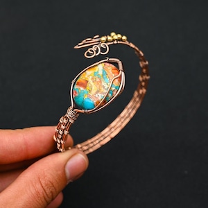 Könnte beinhalten: Ein Armreif aus gewickeltem Kupferdraht mit einem zentralen, ovalen Stein in einem marmorierten Muster aus Türkis, Orange und Weiß. Das Armband besteht aus mehreren Kupferdrahtsträngen mit dekorativen, spiralförmigen Akzenten.
