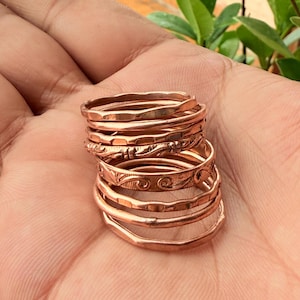 May include: A stack of seven copper rings with varying textures and finishes. The collection includes plain bands, hammered surfaces, and rings featuring engraved floral and swirling patterns, all showcasing the metallic reddish-brown hue of copper.