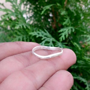 Puede incluir: Un anillo de plata con una banda ondulada e irregular y un acabado martillado y texturizado. La superficie metálica refleja la luz, creando reflejos a lo largo de los bordes curvos de la banda. El anillo tiene una forma orgánica y desigual.