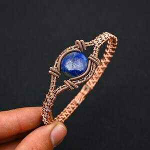 Può includere: Bracciale in filo di rame con una gemma centrale, rotonda, blu scuro. La gemma è incastonata in una cornice di filo di rame intrecciato. Il bracciale ha un design intrecciato ed è esposto su uno sfondo scuro.