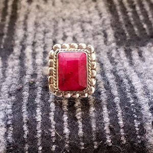 Puede incluir: Un anillo de plata con una gran gema rectangular de color rojo intenso. La gema está engastada en un bisel decorativo y texturizado con un borde en forma de cuerda. El anillo se muestra sobre una superficie gris texturizada.